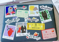 A dark teal laptop covered with colorful, themed stickers including 'Romance', 'Fantasy', 'Fairytale', 'Billionsaire', and 'Age Gap', photographed on a light gray desk.