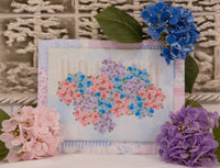 A framed floral needlepoint or cross-stitch design featuring a heart-shaped arrangement of pink, purple, and blue flowers on a light blue background, styled with pink and blue hydrangeas against a textured glass block backdrop.