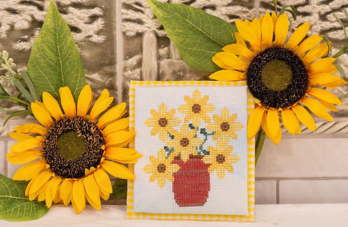 A square cross-stitch sampler depicting a bouquet of yellow sunflowers in a red pot, bordered in yellow gingham, styled with two large artificial sunflowers against a rustic stone wall background.