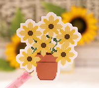 A die-cut sticker featuring a pixelated cross-stitch style design of yellow sunflowers in a terracotta pot, held by a pink pen against a blurred background of sunflowers.