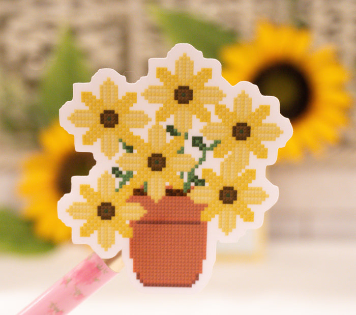 A die-cut sticker featuring a pixelated cross-stitch style design of yellow sunflowers in a terracotta pot, held by a pink pen against a blurred background of sunflowers.