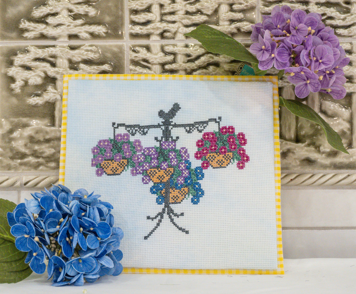 A finished cross-stitch sampler featuring a hanging basket of purple and pink flowers, styled with blue and purple hydrangeas in front of a textured tile background.