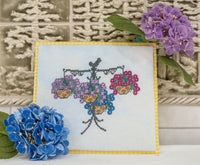 A finished cross-stitch sampler featuring a hanging basket of purple and pink flowers, styled with blue and purple hydrangeas in front of a textured tile background.