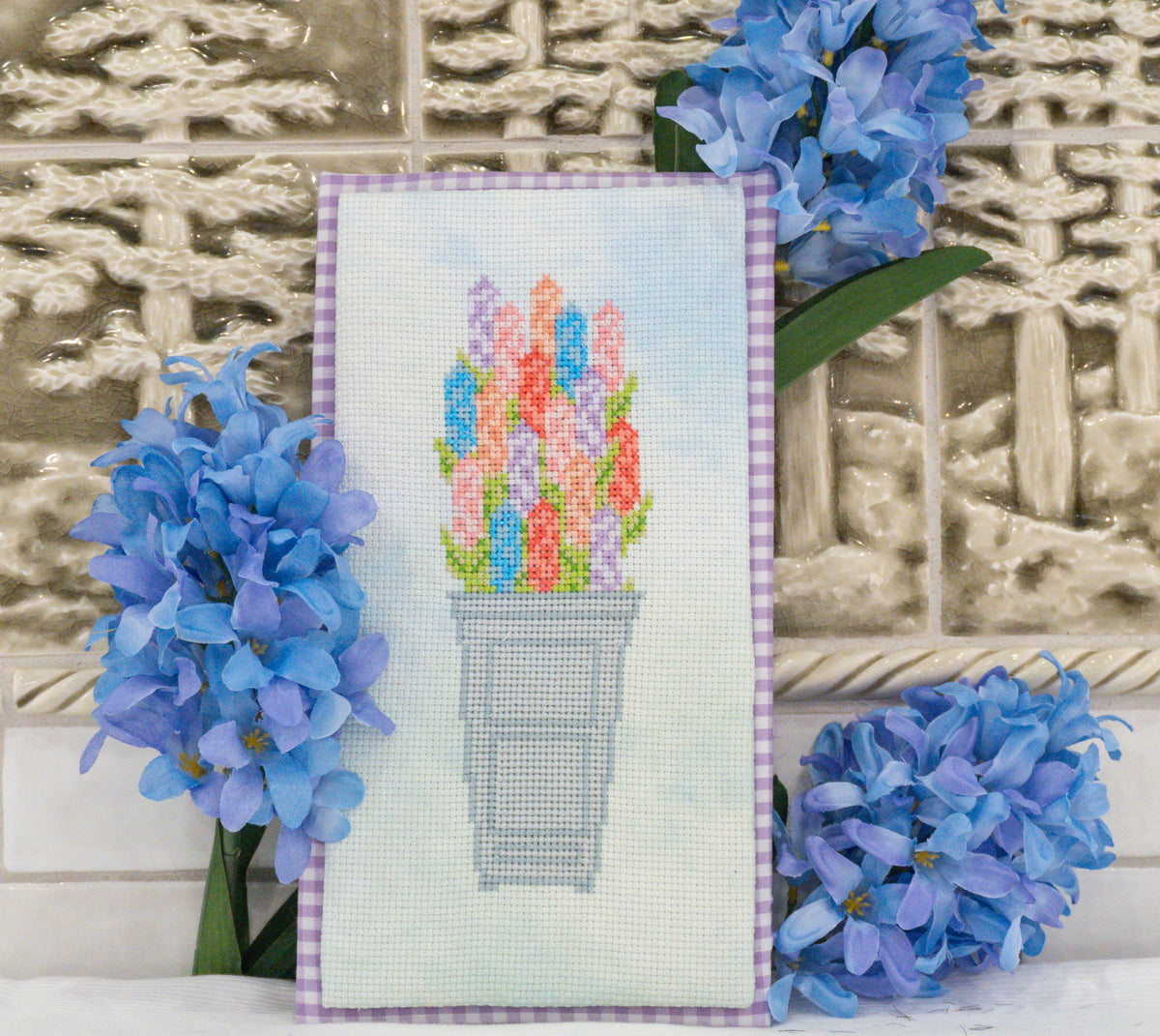 A framed cross-stitch artwork depicting colorful tulips in a gray pot, styled with blue hyacinth flowers against a textured beige tile background.
