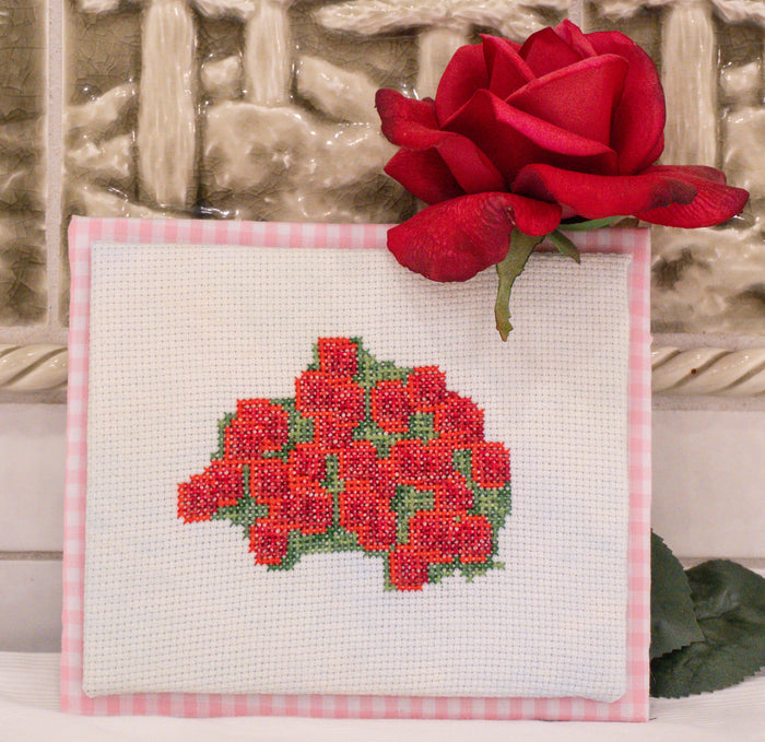 A square white cross-stitch canvas featuring a red rose bouquet with green leaves, bordered by pink gingham fabric, with a real red rose placed on top against a textured beige background.