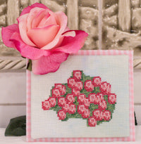 A small cross-stitch or needlepoint canvas featuring a pink and red floral cluster with green leaves on a white grid, bordered by pink gingham. A large pink rose is placed on top, with a decorative textured background behind.