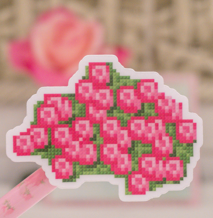 A die-cut sticker featuring a pixel-art style pink rose bouquet with green leaves, shown on a pink ribbon against a softly blurred background.