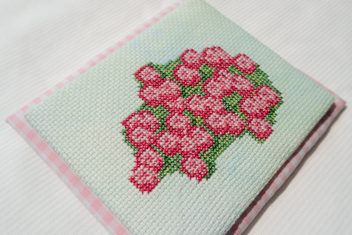 A square embroidered needlepoint canvas featuring a pink rose bouquet on a pale mint green background, finished with a pink and white gingham border, shown on a white textured fabric surface.