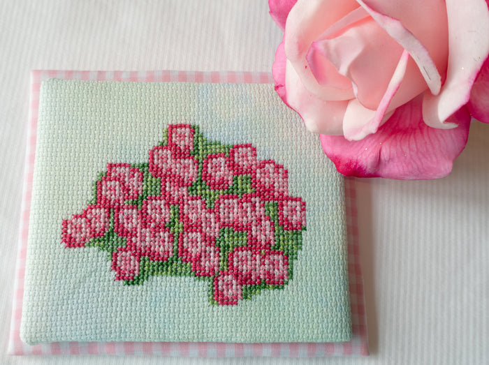A small square cross-stitch piece featuring a pink rose bouquet on a pale mint background, bordered in pink gingham, styled with a real pink rose on a white textured surface.