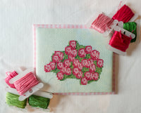 A square cross-stitch sampler featuring a pink rose bouquet on a pale mint background, surrounded by spools of embroidery thread in pink and green on a white fabric surface.