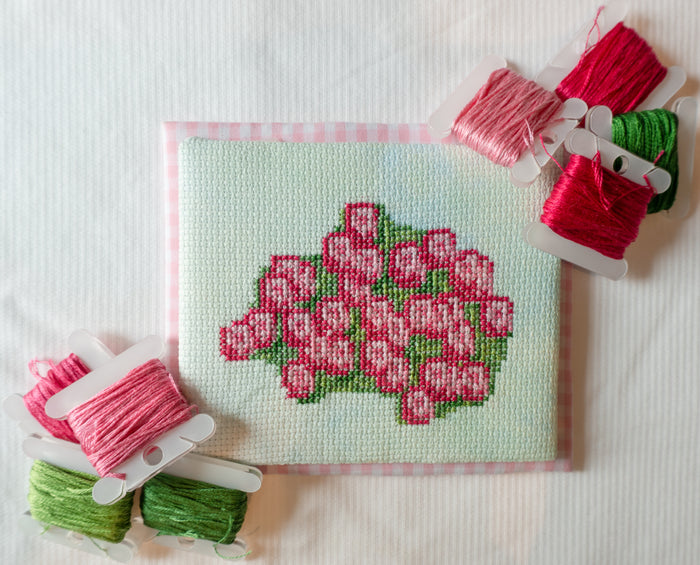 A square cross-stitch sampler featuring a pink rose bouquet on a pale mint background, surrounded by spools of embroidery thread in pink and green on a white fabric surface.
