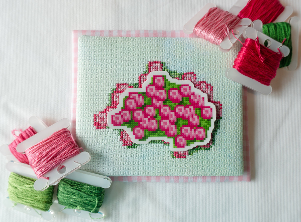 A square embroidery canvas with a pink floral design on a pale green background, surrounded by spools of pink, green, and red thread on a white fabric surface.