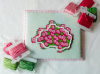 A square embroidery canvas with a pink floral design on a pale green background, surrounded by spools of pink, green, and red thread on a white fabric surface.
