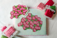 A cross-stitch canvas featuring a pink and green floral bouquet design, surrounded by spools of matching embroidery thread on a white textured fabric background.