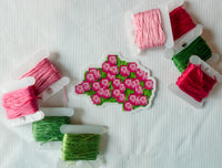 A small embroidered floral patch or applique with pink flowers and green leaves, arranged on a white fabric background with several spools of thread in pink and green.