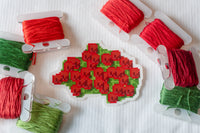 A completed pixel-style cross-stitch design of red berries with green leaves, surrounded by spools of red and green thread on a white fabric background.