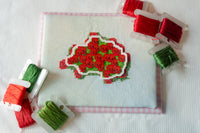 A square white embroidery canvas featuring a stitched red and green red rose bush outline design, surrounded by spools of red and green thread on a white fabric background.