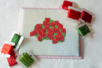 A square white cross-stitch canvas featuring a red floral motif with green leaves, surrounded by spools of embroidery thread in red and green on a white fabric background.