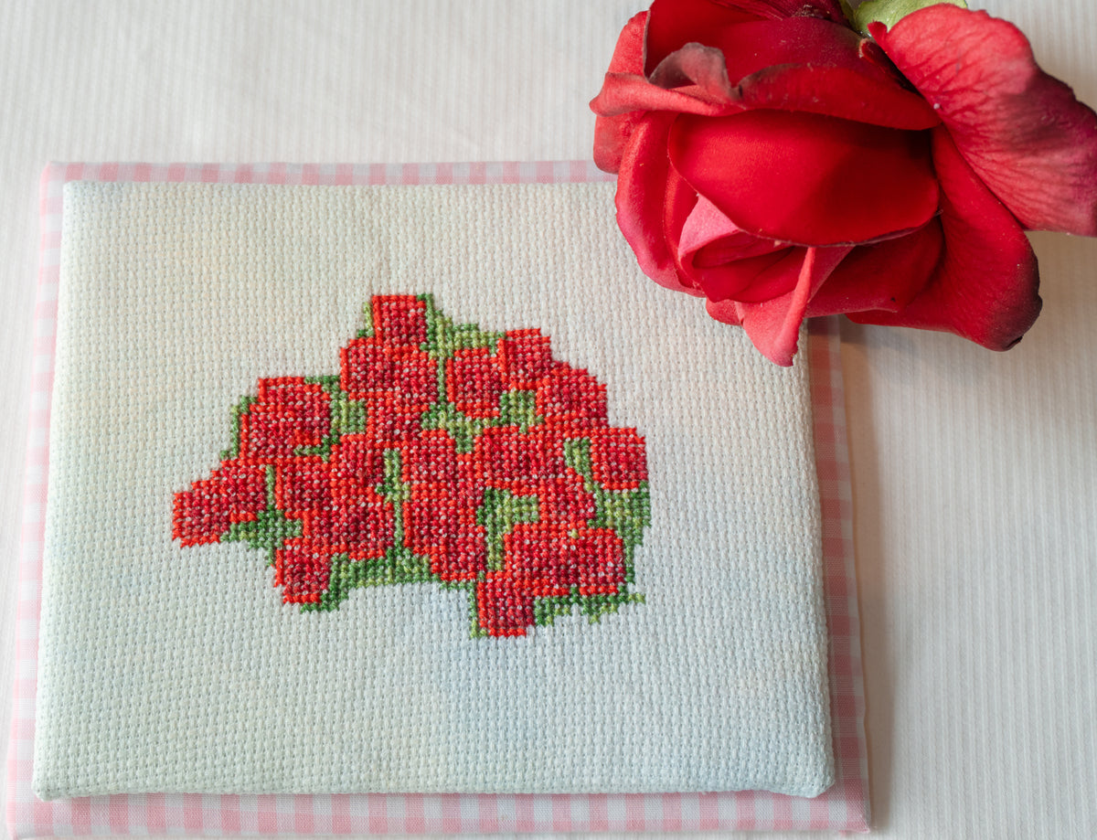 A close-up of a finished cross-stitch piece featuring a red rose bouquet on white A4 canvas, bordered with pink and white gingham fabric, with a real red rose placed on top against a white background.