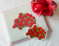 A close-up of a cross-stitch or embroidery sample showing a red rose motif on white fabric, with a matching embroidered patch placed in front and a real red rose resting on top. The background is a white textured surface.