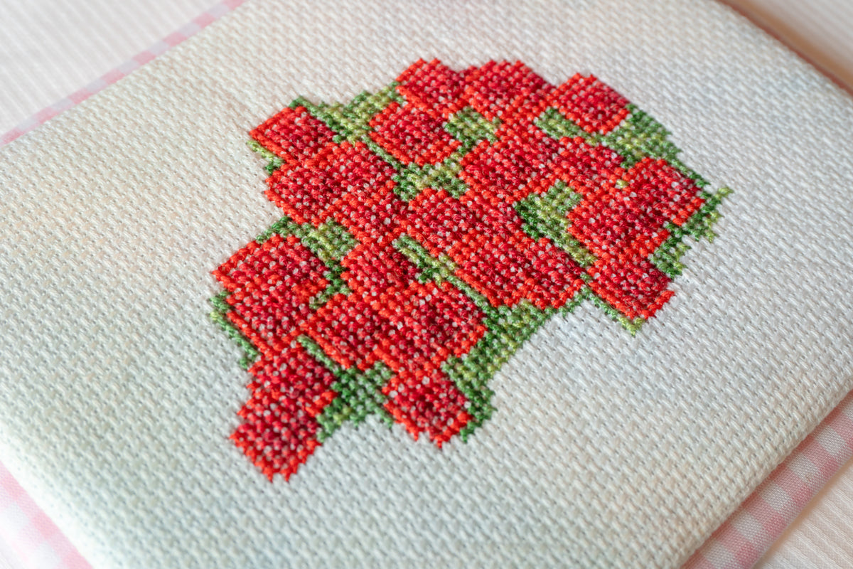 Close-up of a cross-stitch embroidery piece featuring a cluster of red flowers with green leaves stitched on a cream-colored fabric background.