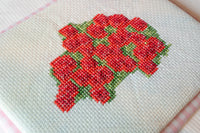 Close-up of a cross-stitch embroidery piece featuring a cluster of red flowers with green leaves stitched on a cream-colored fabric background.
