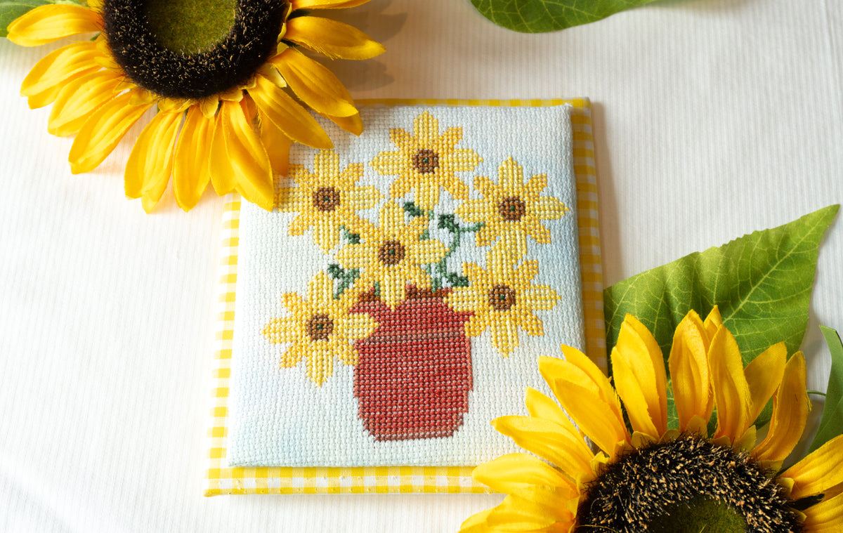 A square cross-stitch or needlepoint piece featuring a bouquet of yellow sunflowers in a red-brown vase on a pale blue background, bordered by yellow-and-white gingham. The piece is styled on a white fabric surface with real sunflowers and green leaves around it.