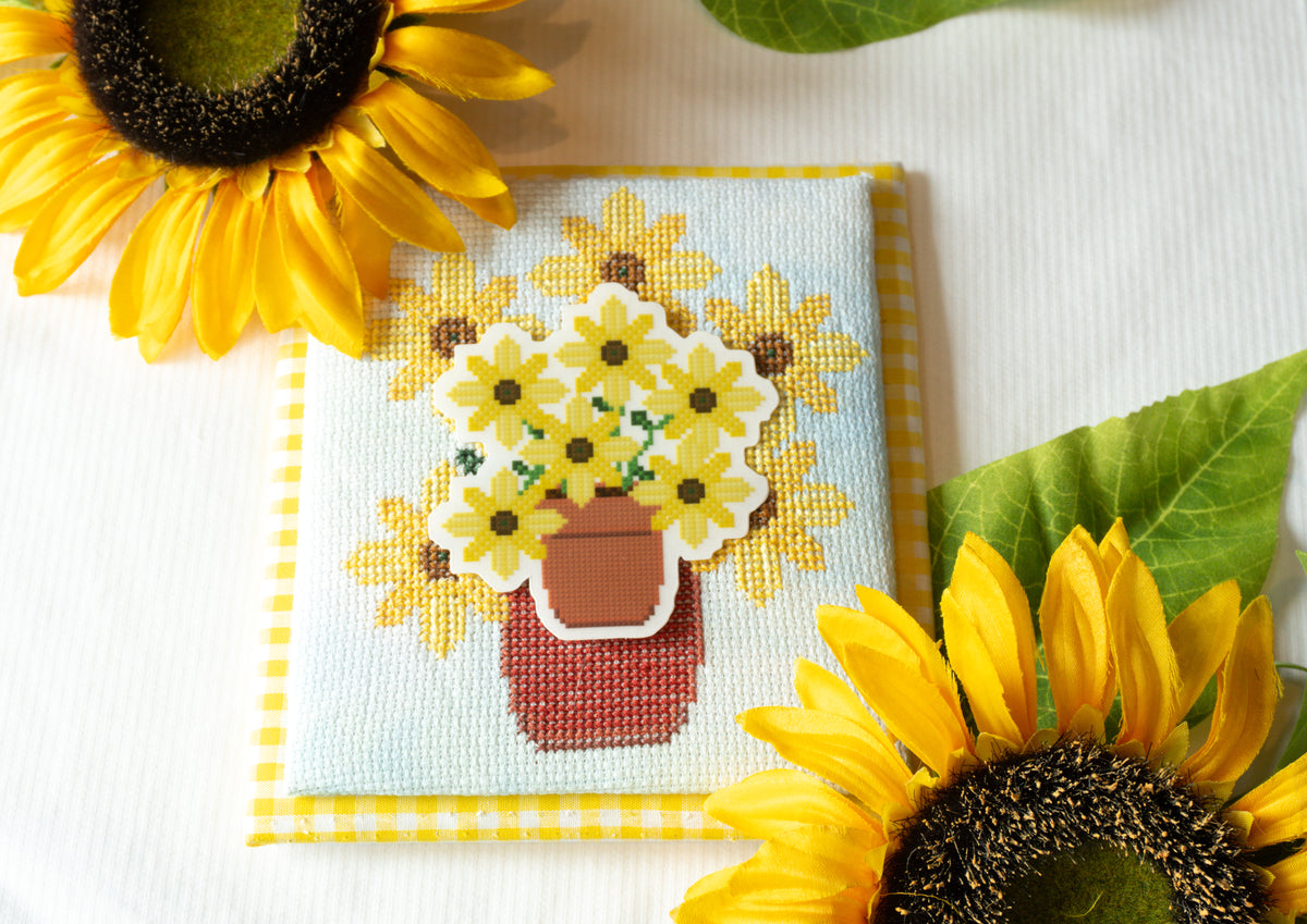 A square cross-stitch sampler featuring a bouquet of yellow sunflowers in a terracotta pot, styled on a white textured fabric with real sunflowers and green leaves.
