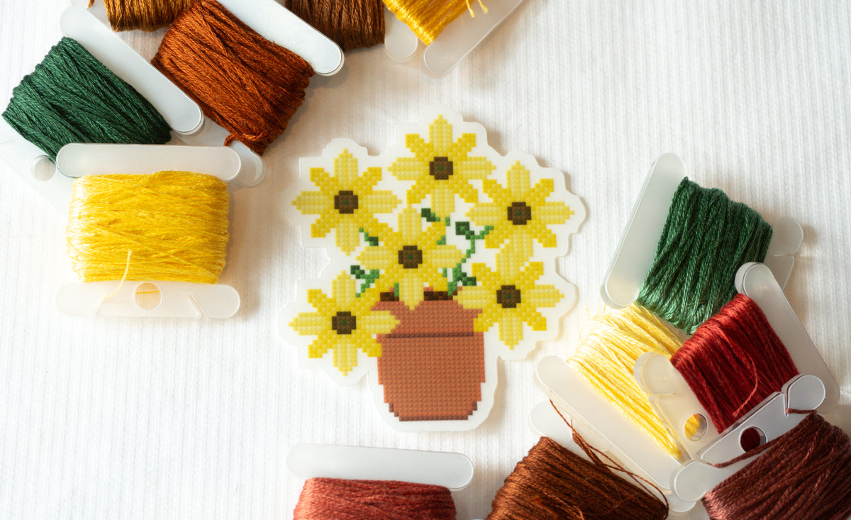 A cross-stitch sampler depicting a bouquet of yellow sunflowers in a brown pot, surrounded by spools of embroidery thread in yellow, green, brown, and red on a white textured background.
