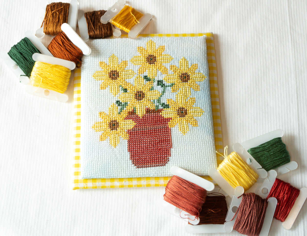 A square cross-stitch sampler featuring yellow sunflowers in a red pot on a light blue background, surrounded by spools of embroidery thread on a white fabric surface.