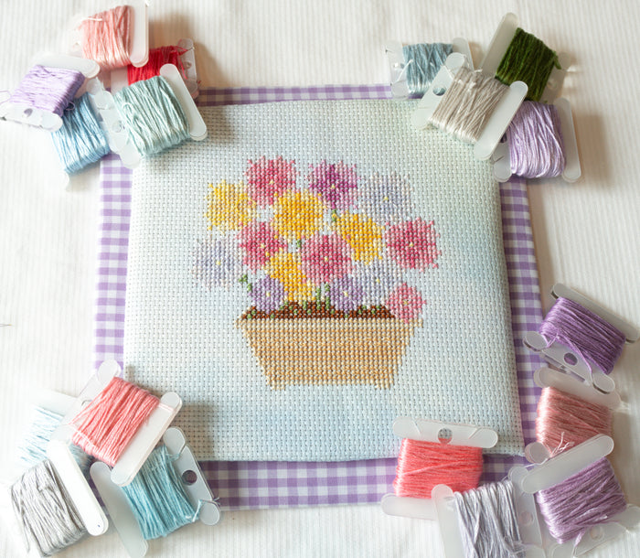 A completed cross-stitch design of a flower pot with colorful flowers, surrounded by spools of thread in various colors on a white fabric background.