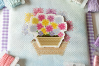 A close-up of a cross-stitch canvas featuring a colorful flower pot design, surrounded by spools of thread in pastel colors on a light blue A4 fabric background.