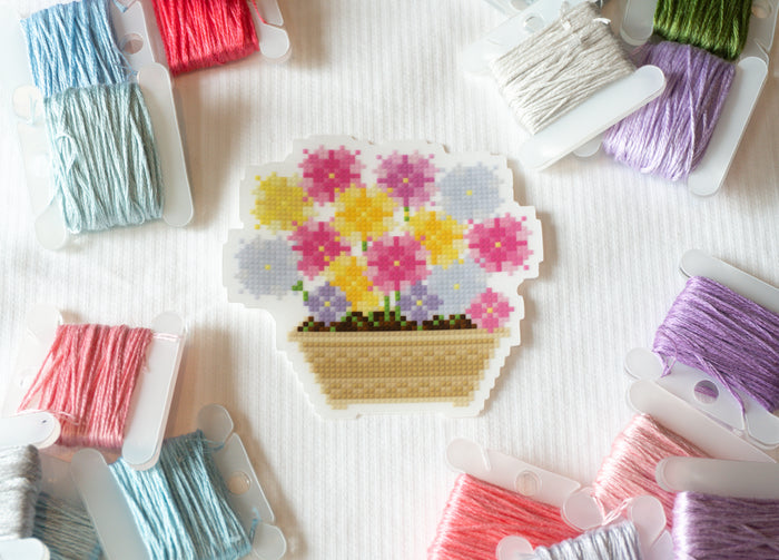 A cross-stitch sampler depicting a basket of colorful flowers, surrounded by various spools of embroidery floss in pastel and bright colors on a white textured fabric background.