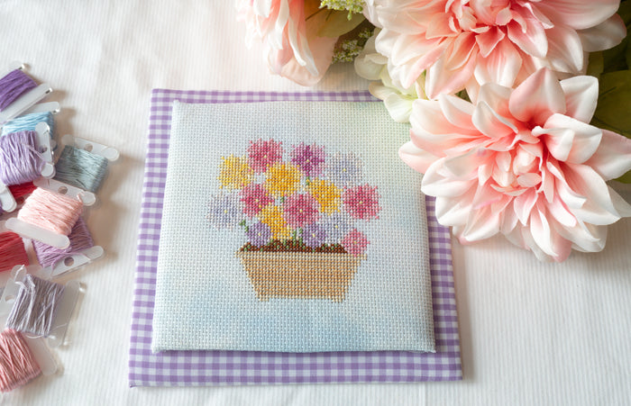 A square cross-stitch sampler featuring a colorful flower pot with pink, yellow, and purple blossoms on a light blue background, placed on a purple-and-white gingham mat. The sampler is styled on a white fabric surface with pink flowers and spools of thread nearby.