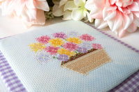 A close-up of a completed cross-stitch design featuring a basket of colorful flowers on a light blue fabric background, styled with pink and white flowers and a purple gingham cloth.