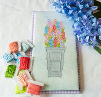 A completed cross-stitch design of colorful flowers in a pot, displayed on a white fabric background with loose spools of embroidery thread and blue flowers nearby.