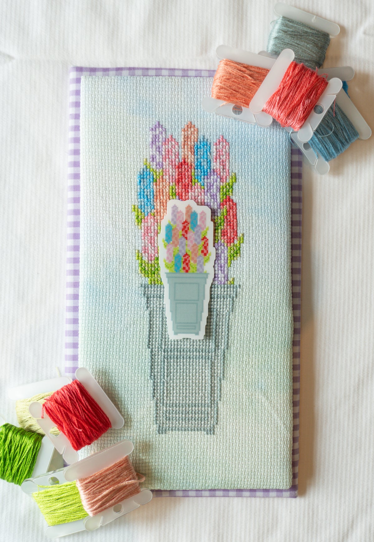 A completed cross-stitch embroidery piece depicting colorful hyacinths in a light blue-gray pot, displayed on a white fabric background with several spools of matching embroidery thread arranged around it.