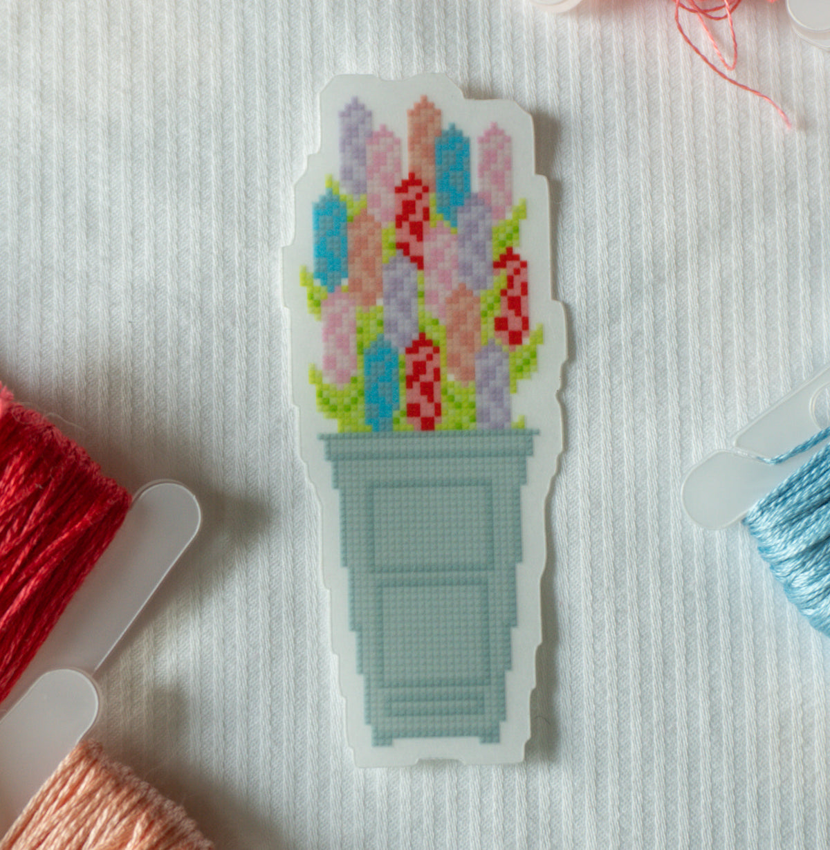 A die-cut sticker featuring a pixel-art style cross-stitch design of colorful flowers in a light blue-gray pot, placed on a white textured fabric background with spools of thread partially visible around it.