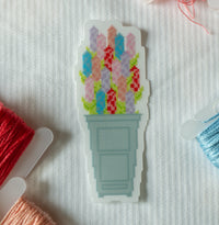 A die-cut sticker featuring a pixel-art style cross-stitch design of colorful flowers in a light blue-gray pot, placed on a white textured fabric background with spools of thread partially visible around it.
