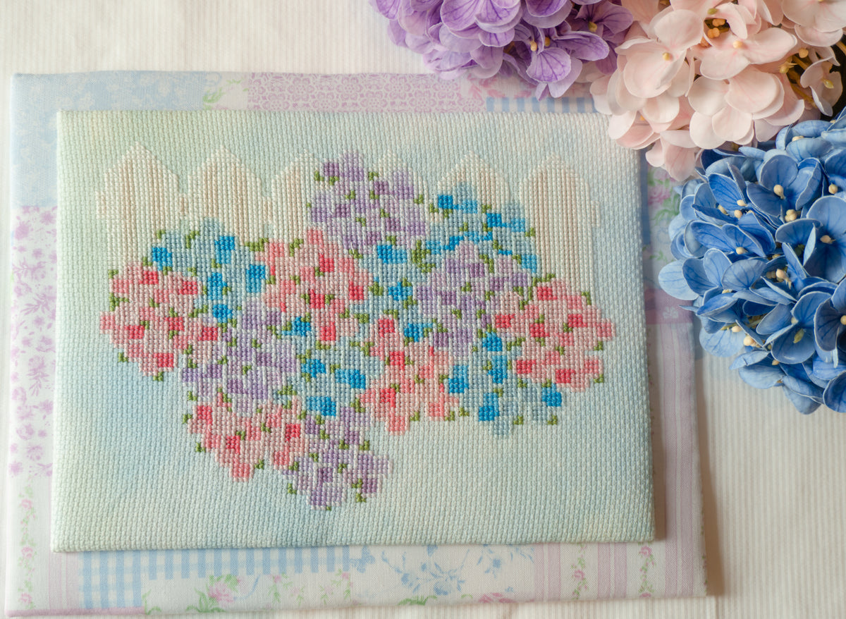 A square embroidered canvas featuring a pastel floral heart design with pink, purple, and blue flowers on a pale blue background, styled with decorative flowers in the corner.