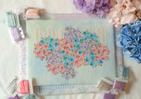 A completed cross-stitch canvas featuring a heart-shaped floral pattern in pastel pink, purple, and blue, surrounded by spools of matching embroidery thread and decorative flowers on a white fabric background.