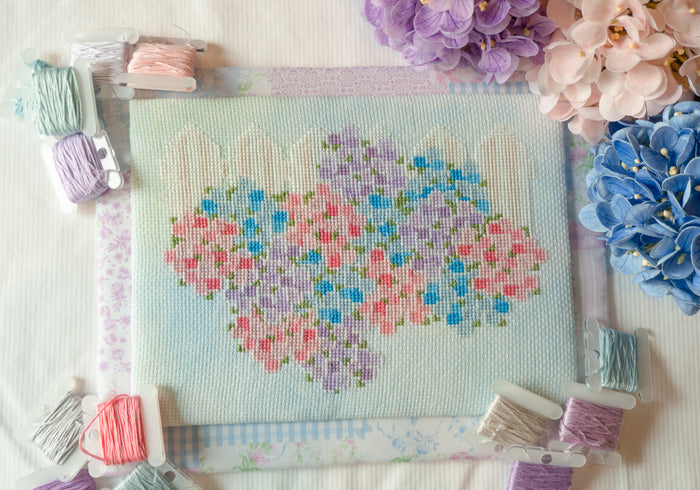 A completed cross-stitch canvas featuring a heart-shaped floral pattern in pastel pink, purple, and blue, surrounded by spools of matching embroidery thread and decorative flowers on a white fabric background.