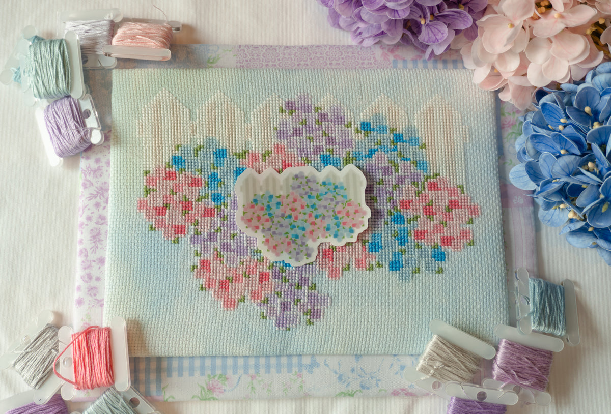 A floral cross-stitchcanvas with embroidered pink, purple, and blue flowers, surrounded by spools of thread and decorative flowers on a white fabric background.