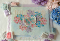 A floral cross-stitchcanvas with embroidered pink, purple, and blue flowers, surrounded by spools of thread and decorative flowers on a white fabric background.