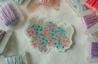 A scalloped white sticker or patch featuring a colorful floral pattern of pink, blue, and purple flowers with green leaves, displayed on a white textured fabric surrounded by spools of embroidery thread.
