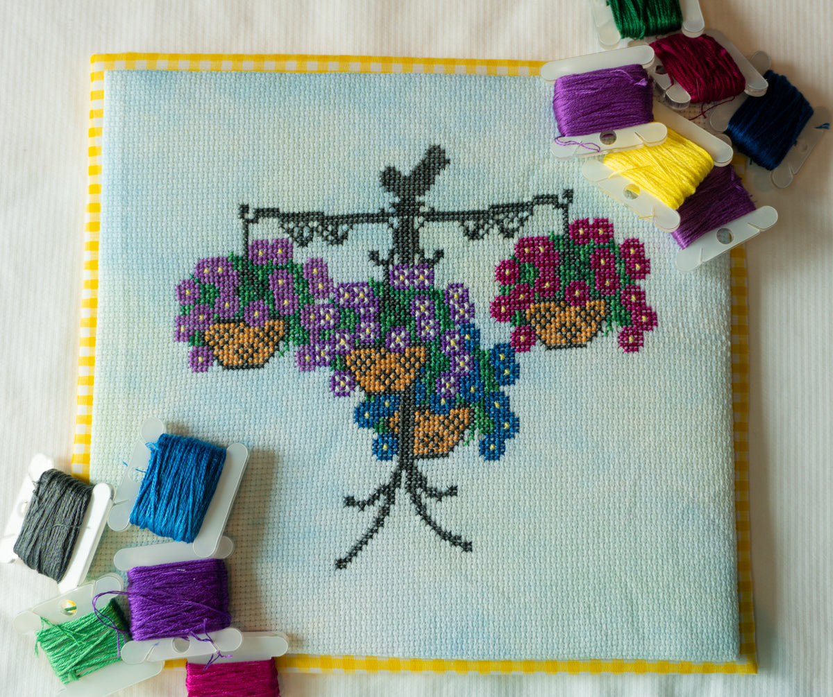 A completed cross-stitch piece featuring a black tree with purple, pink, blue, and yellow flowers in hanging baskets on a light blue fabric background, bordered in yellow. Multiple spools of thread in various colors are arranged around it on a white fabric surface.