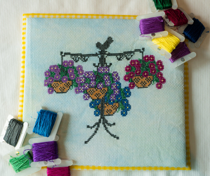 A completed cross-stitch piece featuring a black tree with purple, pink, blue, and yellow flowers in hanging baskets on a light blue fabric background, bordered in yellow. Multiple spools of thread in various colors are arranged around it on a white fabric surface.