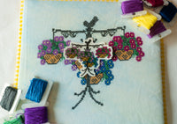 A close-up of a cross-stitch canvas featuring a decorative floral and basket design on a pale blue fabric, surrounded by spools of colorful thread on a white surface.
