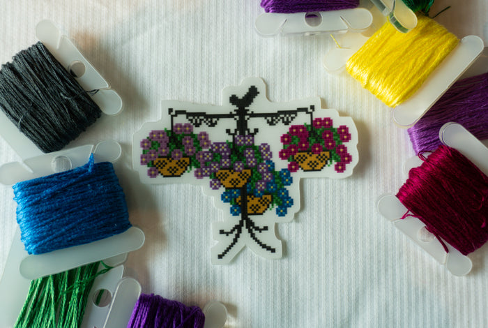 A cross-stitch embroidery sample depicting hanging baskets of purple and blue flowers with yellow centers, surrounded by spools of multicolored thread on a white textured fabric background.