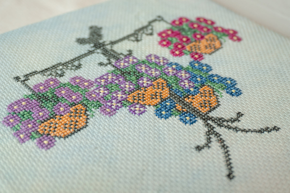 Close-up of a cross-stitch embroidery on light blue fabric, featuring purple, pink, orange, and blue flowers with green leaves and black branch outlines.